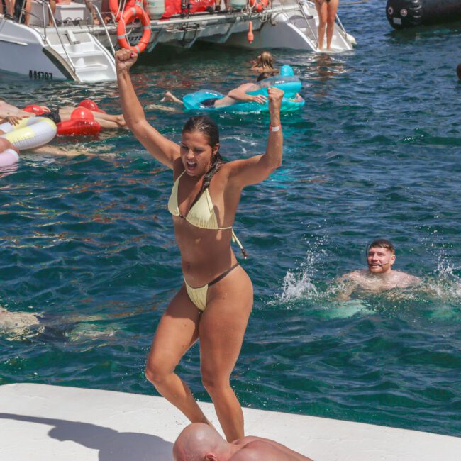 A woman in a yellow bikini stands and raises her arms triumphantly on a floating platform in the water, surrounded by people swimming and relaxing on inflatables near anchored boats.