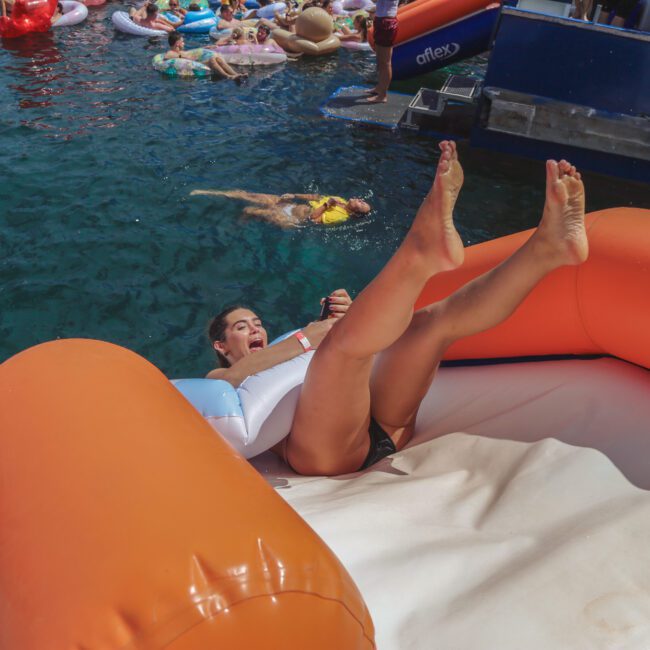 A person slides down an orange inflatable slide into a body of water, holding a white float. Others relax on various inflatables in the water nearby, enjoying a sunny, fun-filled day.