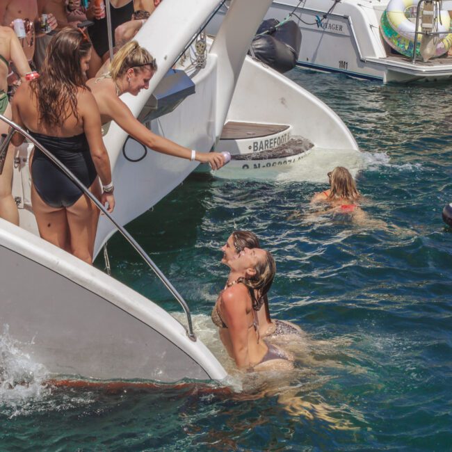 Several women in swimsuits enjoy a party on and around a yacht, some climbing out of the water and others standing on the boat’s deck, with another boat and floating toys visible in the background.