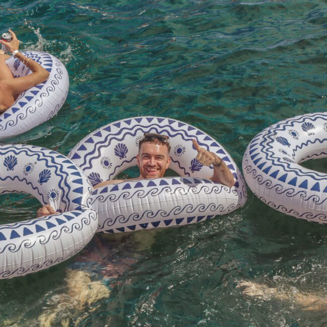 A man smiles and poses with his arms through two inflatable pool rings in clear blue water, surrounded by others on similar floats. The scene is relaxed and cheerful. The "Yacht Social" logo is in the bottom right corner.