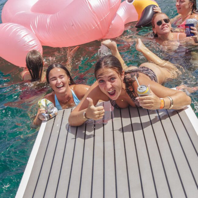 A group of young women enjoy a sunny day in the water near a dock, holding drinks and smiling. One woman gives a thumbs up while lying on the dock, with an inflatable pink float visible behind them.
