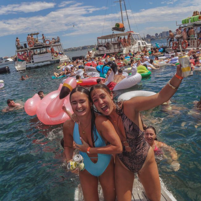 Two women in swimsuits smile and pose on a float in the ocean, surrounded by people on inflatables and boats, enjoying a lively party under a sunny sky. Many hold drinks and there are various colorful pool floats.
