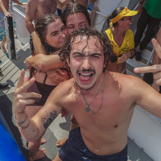 A young man in swimwear poses enthusiastically on a boat, making hand signs and smiling. Behind him, several people in swimsuits enjoy the sunny day, some hugging and laughing. The sea is visible beside the boat.