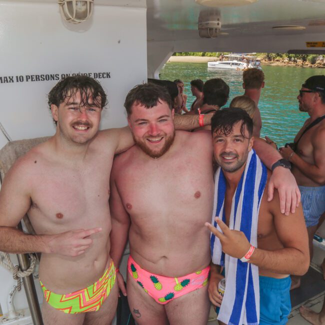 Three smiling men in colorful swim trunks pose together on a crowded boat. The group is enjoying a sunny day, with turquoise water and other people in swimsuits visible in the background.