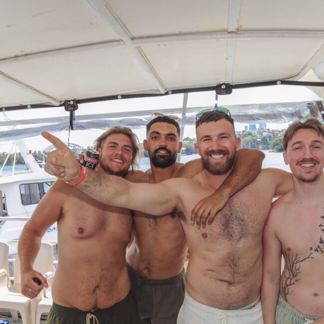Four smiling shirtless men stand close together on a boat, with arms around each other. The man on the left points outward. Other people and water are visible in the background. The scene is bright and festive.