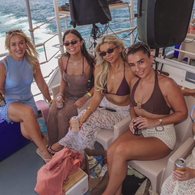 Five women in swimwear and sunglasses sit together on a boat, smiling and holding drinks. The ocean and boat railing are visible in the background, suggesting a sunny, relaxed atmosphere.