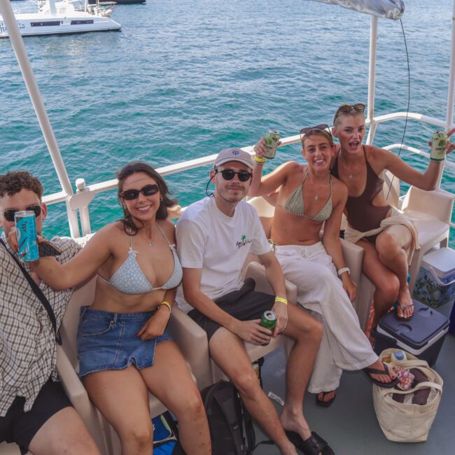Five friends sit and smile on a boat, holding drinks and wearing summer clothes and swimsuits. The ocean and other boats are visible in the background on a sunny day.