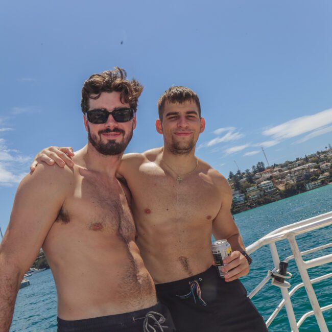 Two shirtless men stand smiling and posing together on a sunny boat ride, with one arm around each other. Behind them are blue waters, a city shoreline, and a clear sky.