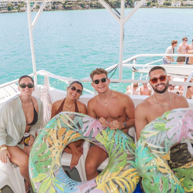 Four smiling people in swimwear sit on a boat holding colorful inflatable tubes, with bright blue water and other people on deck in the background. It’s a sunny day and everyone looks relaxed and happy.
