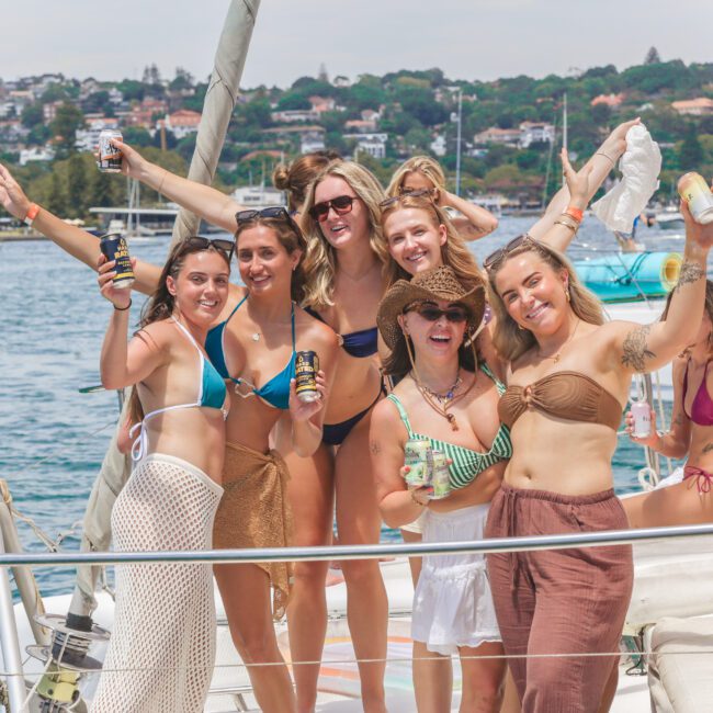 A group of six women in swimsuits pose and smile on a boat, holding drinks, with water, yachts, and a shoreline with houses visible in the background on a sunny day.