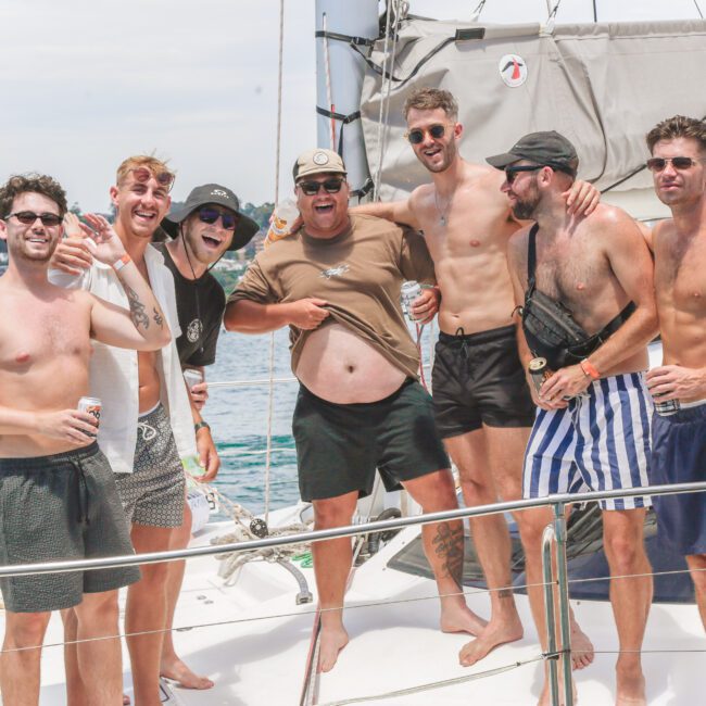 Seven smiling men, most shirtless, pose together on a boat in the sun. One man lifts his shirt to show his stomach, while others hold drinks. The background features water, other people, and shoreline greenery.