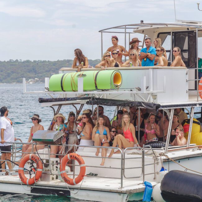 A group of people in swimsuits relax and enjoy drinks on a two-story boat with inflatable pool floats. The boat is on the water near a forested shoreline under a partly cloudy sky.