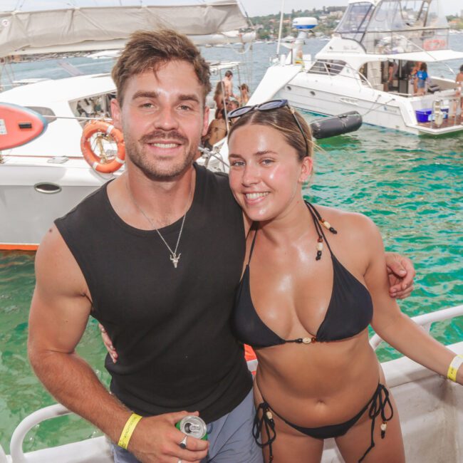 A man and woman in swimwear smile for a photo on a boat, with yachts and people enjoying the water in the sunny background. The woman holds a blue cup, and both have event wristbands.