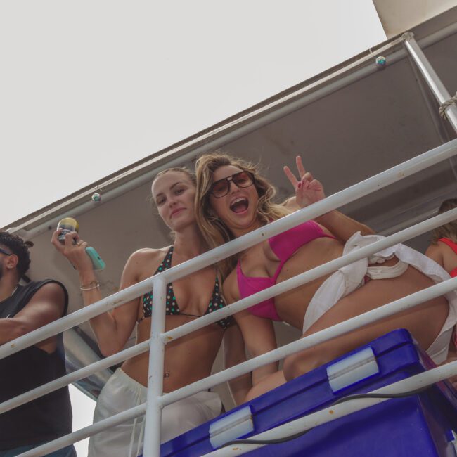 Three people in swimwear stand on a metal balcony; two women in bikinis smile and pose, one flashing a peace sign, while a bearded man in sunglasses stands nearby. The scene appears festive and summery.