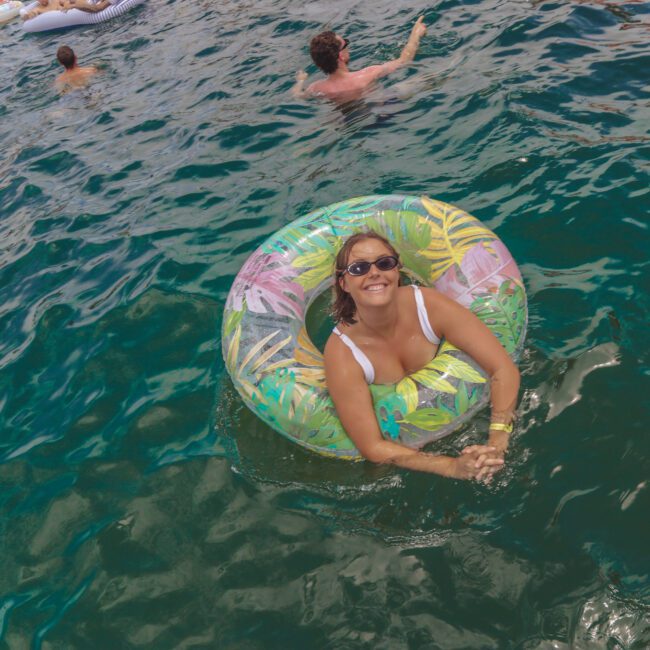 A smiling woman wearing sunglasses floats on a colorful inflatable ring in clear blue-green water, surrounded by other people enjoying the water in the background.