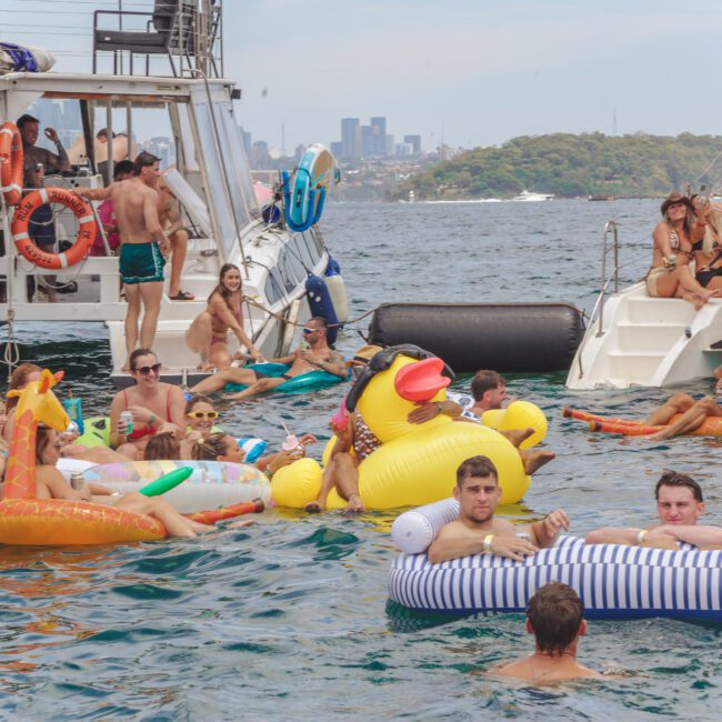A group of people relax on colorful inflatable pool floats between two boats on a sunny day, with a city skyline and trees in the background. Some are in the water, while others are sitting or standing on the boats.