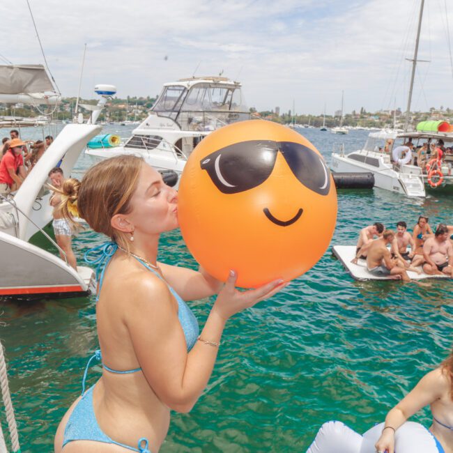 A woman in a blue bikini kisses a large orange beach ball with sunglasses printed on it at a boat party. Several people in swimwear relax on boats and floating platforms on a sunny day.
