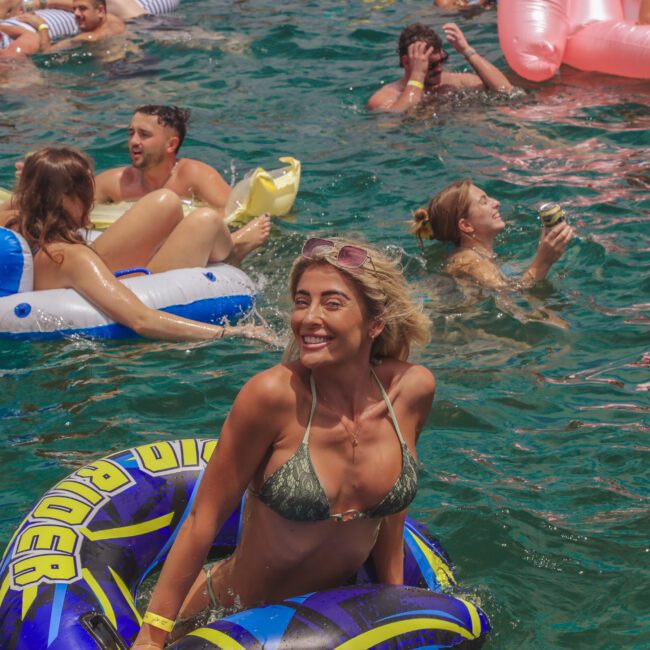 A woman in a green bikini smiles while floating in a blue inflatable tube on the water, surrounded by other people swimming and relaxing on pool floats during a sunny outdoor party.
