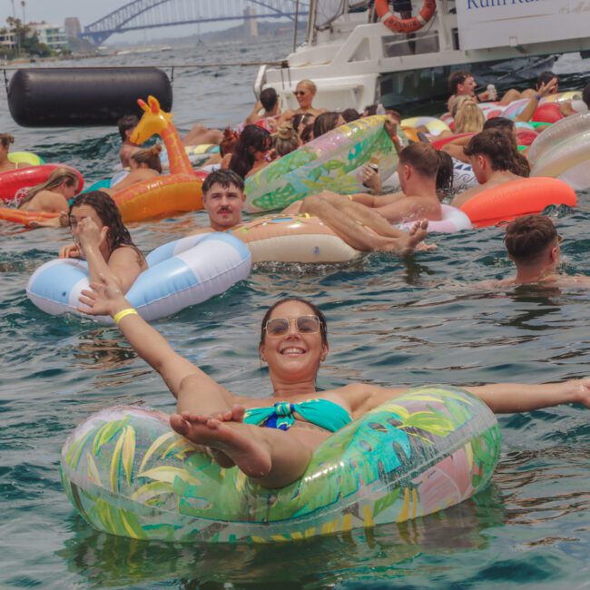A woman in sunglasses smiles while relaxing on a green inflatable ring in the water, surrounded by people on colorful floaties during a lively boat party. A bridge and boats are visible in the background.