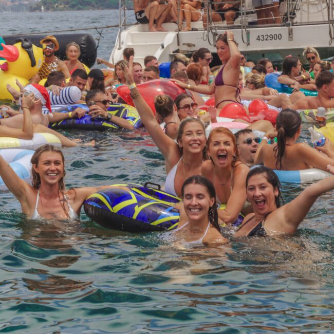 A large group of people enjoy a lively pool party on the water, smiling and waving while floating on colorful inflatables near boats on a sunny day.