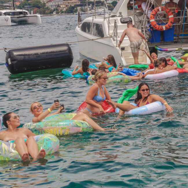 A group of people relax on colorful inflatable floats in the water near anchored boats, smiling and enjoying themselves. Other people are on the boats in swimsuits, with a scenic waterfront in the background.