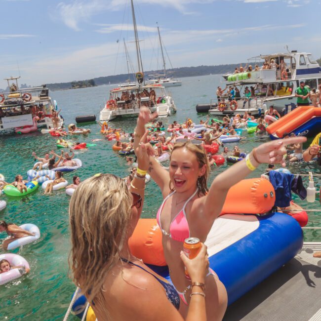 A lively party scene on the water, with people in swimsuits on floaties and boats. Two women in the foreground celebrate on an inflatable, while others relax and dance in the sun. Several boats are anchored nearby.