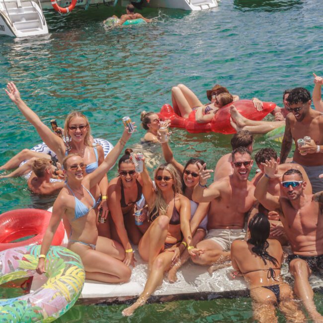 A group of people in swimsuits relax and smile on pool inflatables and a floating mat in clear blue water, holding drinks and waving with boats visible in the background.