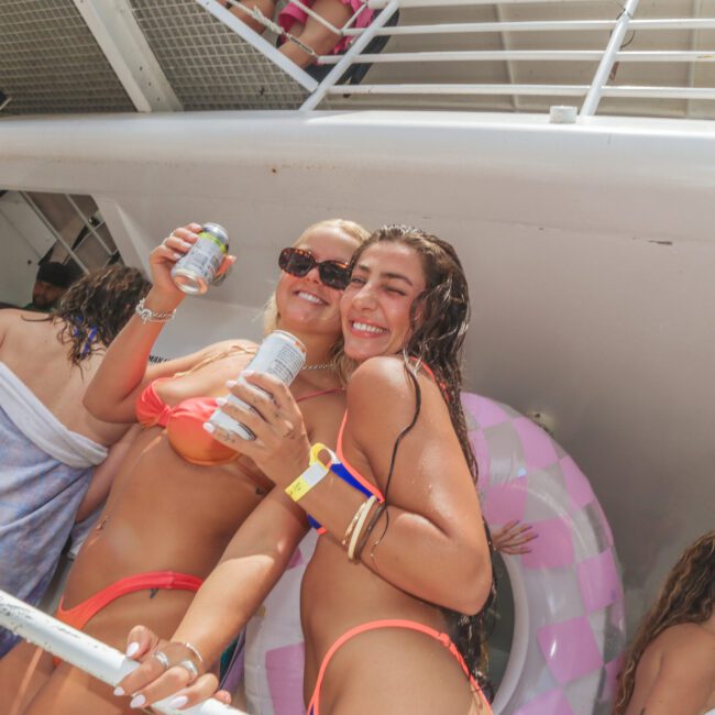 Two women in swimsuits smile and pose with drinks on a crowded boat, surrounded by people and pool floats, enjoying a sunny day.