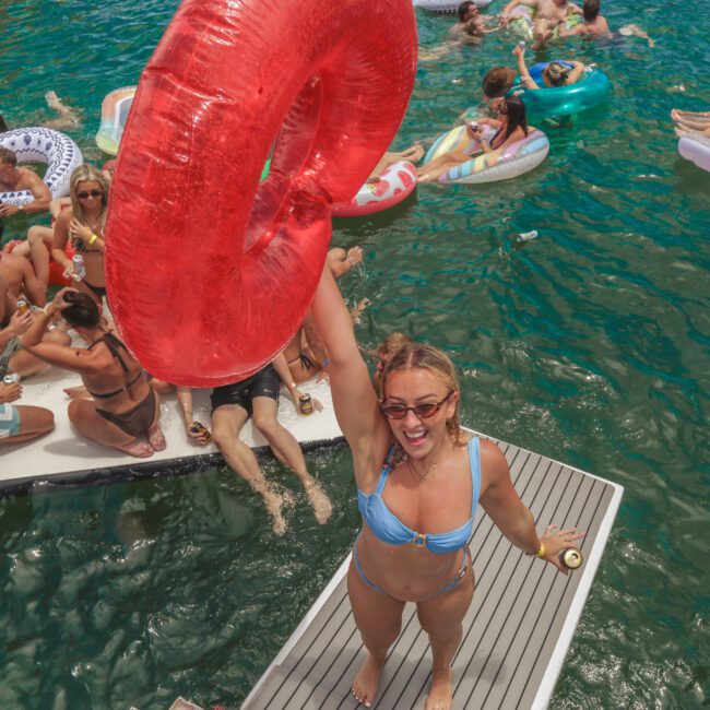 A woman in a blue bikini holds up a large red pool float while smiling on a dock, surrounded by people relaxing on inflatable floats and paddle boards in clear, green water. The scene is lively and festive.