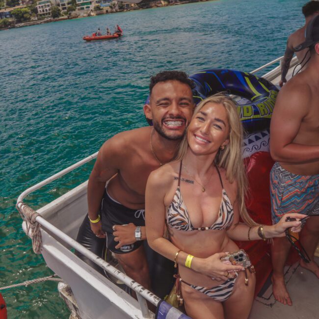 A smiling man and woman in swimsuits pose together on a boat with turquoise water in the background. Other people are around them, enjoying drinks and the sunny weather. A small boat is visible on the water behind them.