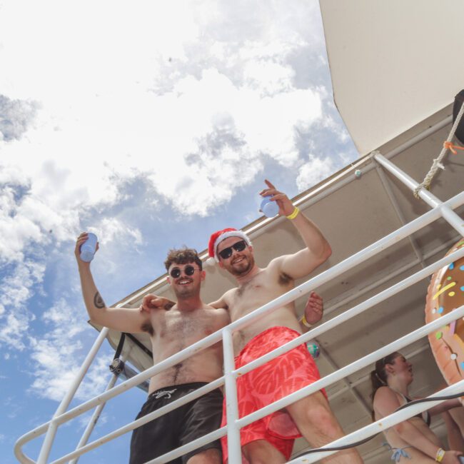 Two men in swim trunks and sunglasses smile and pose on a boat deck under a partly cloudy sky. A donut-shaped pool float is visible on the railing, and other people are in the background enjoying the boat.