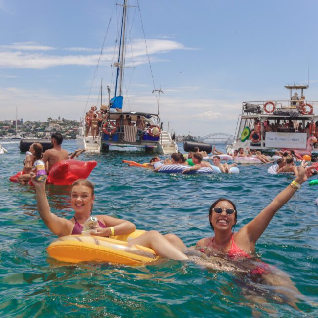 Two people smile and wave while floating on inflatable loungers in blue water, surrounded by others on floats and boats at a lively party. City buildings and the Sydney Harbour Bridge are visible in the background.