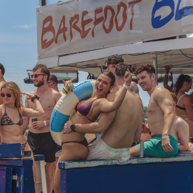 A group of young adults in swimwear enjoy a lively boat party. Two people sit smiling with pool floats, while others stand and chat in the background under a sign reading “Barefoot.” It’s sunny and festive.