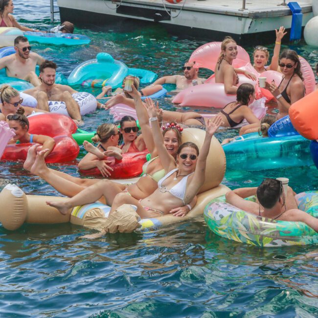 A group of young adults relax and have fun on colorful inflatable floats in the water near a dock; they are smiling, laughing, and enjoying a sunny day together.