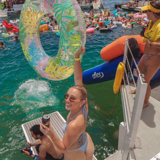 A woman in a bikini smiles at the camera, holding a colorful inflatable ring above her head on a boat deck. People swim and relax on pool floats in the water, with boats anchored nearby under a sunny sky.