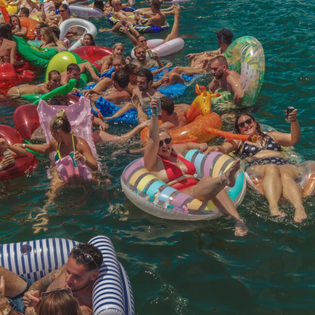 A group of people relax and have fun on colorful inflatable pool floats in blue water, holding drinks and smiling on a sunny day at a lively outdoor party.