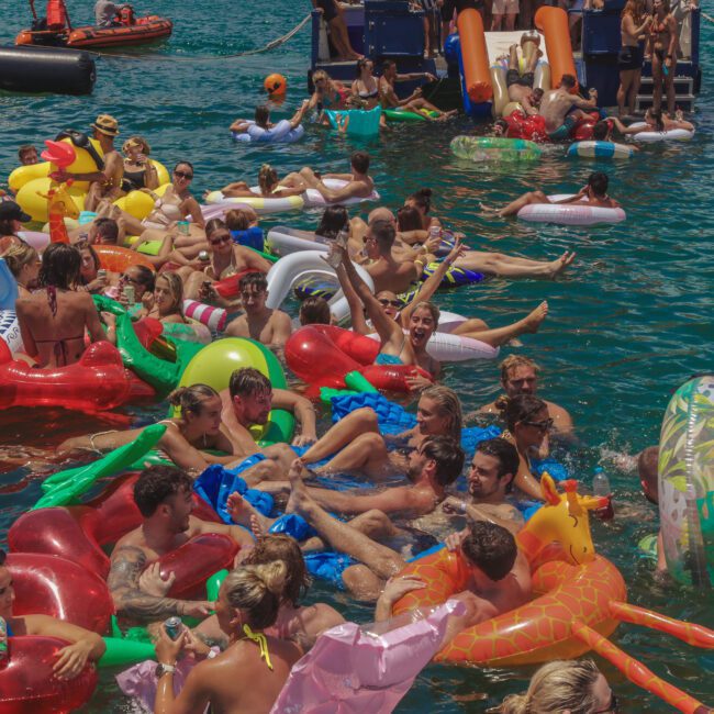 A large group of people relaxing and socializing on colorful inflatable pool floats in the water during a lively summer party, with boats in the background and many attendees holding drinks.