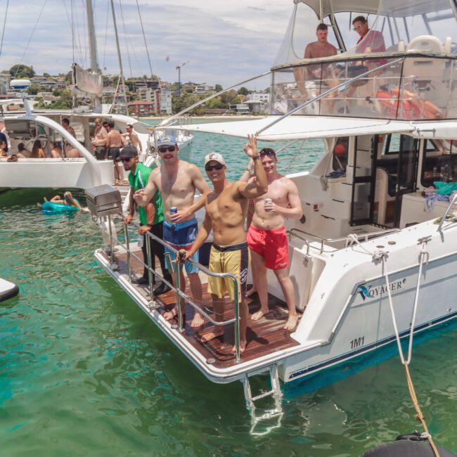 A group of people in swimsuits relax and wave from the back deck of a white yacht on turquoise water, with others on nearby boats enjoying a sunny day. City buildings and greenery are visible in the background.
