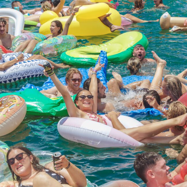A large group of people enjoy a sunny day on colorful pool floats in crowded blue water, smiling and raising drinks, creating a lively, festive atmosphere.