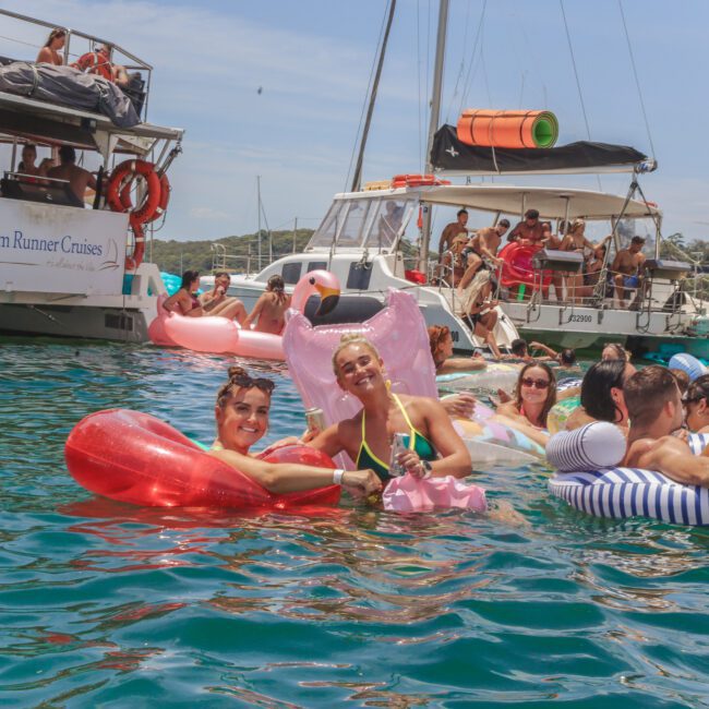 A group of people relax on colorful pool floats in the water near anchored boats during a sunny day. Some smile at the camera, holding drinks, while others chat nearby. The scene is lively and festive.