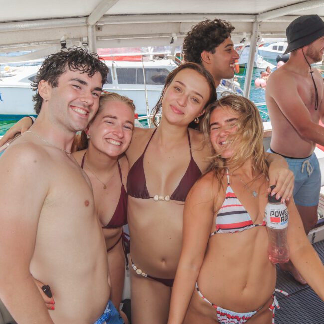 Four young adults in swimsuits smile and pose together on a boat during a sunny day, with other people and boats visible in the background. One person holds a drink and everyone appears to be enjoying a lively social event.