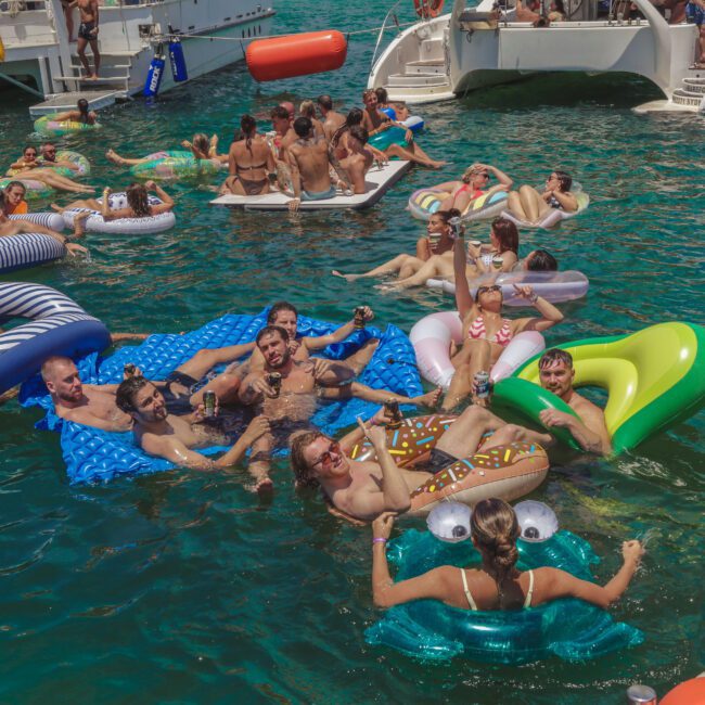 A group of people relax and have fun on colorful inflatable floats in the water near boats on a sunny day, enjoying a lively party atmosphere.