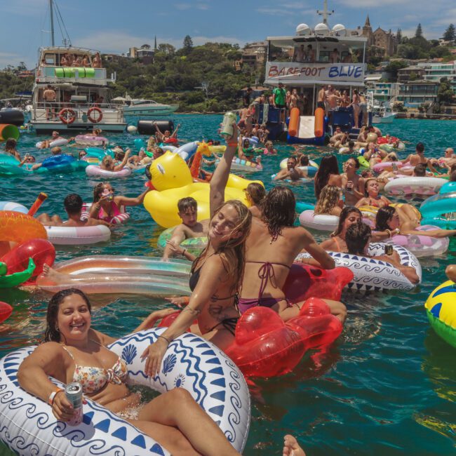 A large group of people on colorful inflatable floats enjoying a sunny day in the water near boats. Many are smiling, waving, or holding drinks, with houses and greenery visible in the background.