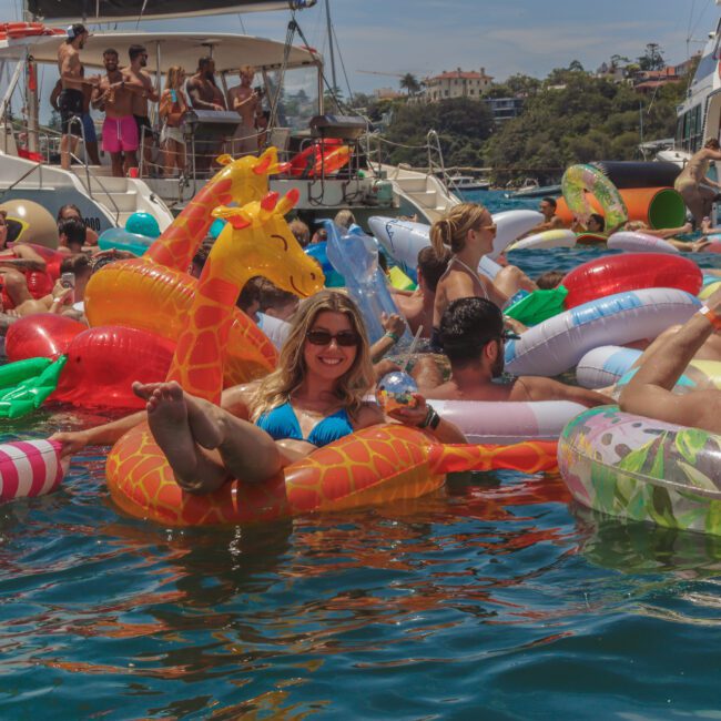 People float in a crowded bay on colorful inflatable pool toys, including a large giraffe. Yachts are anchored nearby, and the atmosphere is festive and lively under sunny skies.
