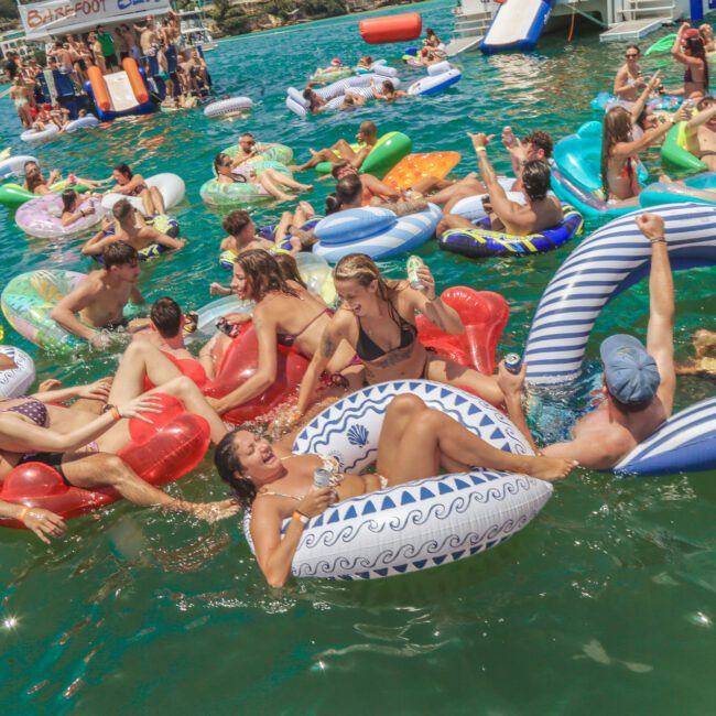 A lively group of people float on colorful inflatables and interact in the water at a crowded outdoor pool party, with boats and more inflatables in the background under a sunny sky.