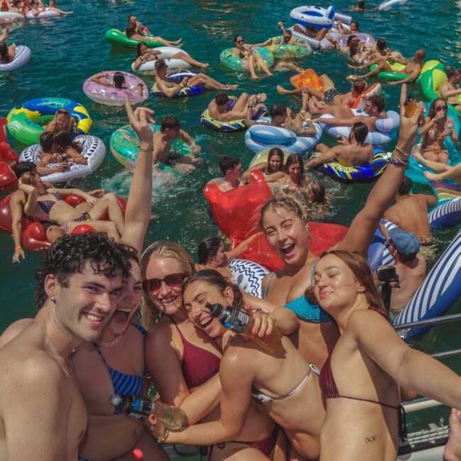 A large group of people in swimsuits enjoy a lively pool party, lounging on colorful inflatables and posing happily for the camera while gathered closely on a boat and in the water, surrounded by sunshine and blue water.