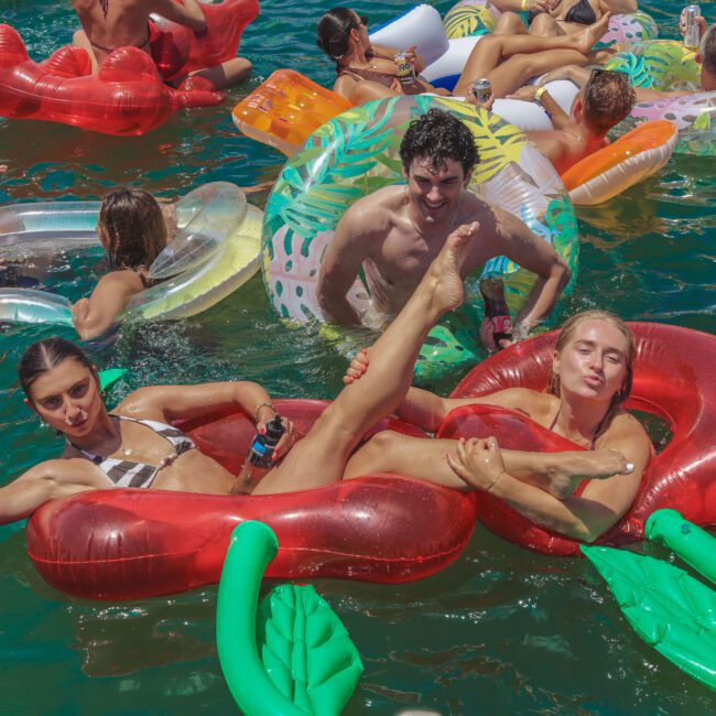 A group of young people relax on colorful inflatable pool floats in sunny water. Two women pose playfully on a cherry-shaped float, while others smile and enjoy drinks nearby. The mood is lively and fun.