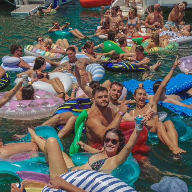 A large group of people in swimsuits relax and have fun on colorful inflatable pool floats in a lake, raising their drinks and smiling at the camera on a sunny day. The atmosphere is lively and festive.