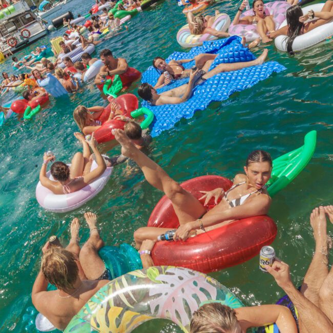 People relax on colorful inflatables in a crowded, lively lake party. Many are holding drinks and chatting under the sun, with boats and more floaties in the background. The water is bright blue and the atmosphere is festive.