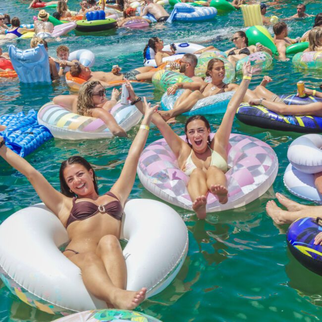 A large group of people relax and have fun on colorful inflatable pool floats in a sunny, crowded body of water. Two women in the foreground smile and raise their arms, enjoying the lively summer atmosphere.